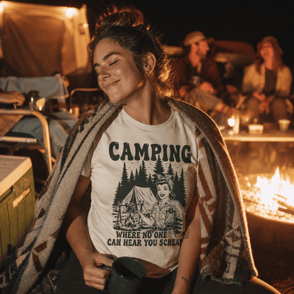 A woman sits by a campfire, wearing a graphic t-shirt that reads "CAMPING WHERE NO ONE CAN HEAR YOU SCREAM" with a retro design.