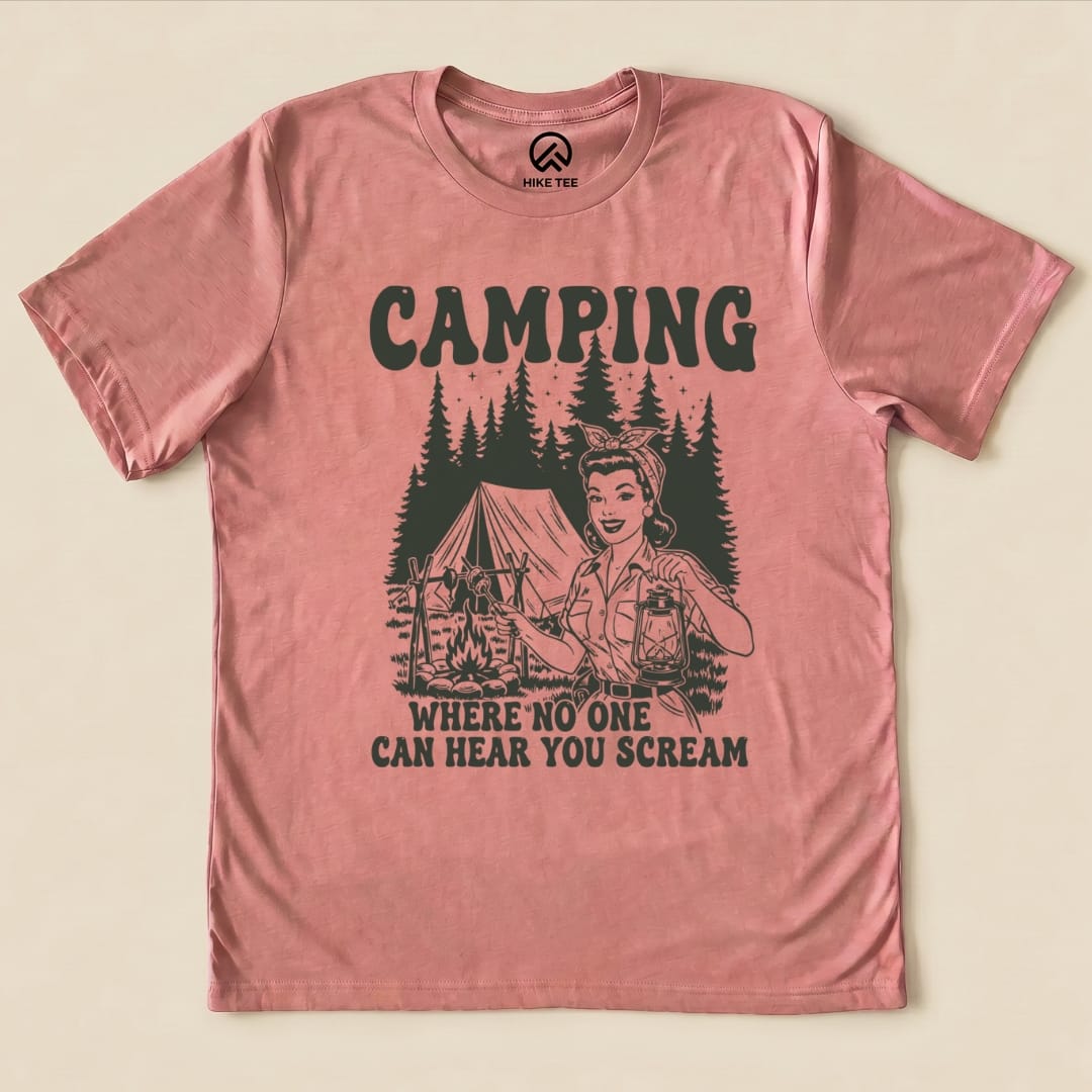 A graphic t-shirt featuring a retro design with the text "CAMPING WHERE NO ONE CAN HEAR YOU SCREAM" and an illustration of a woman holding a lantern near a tent.