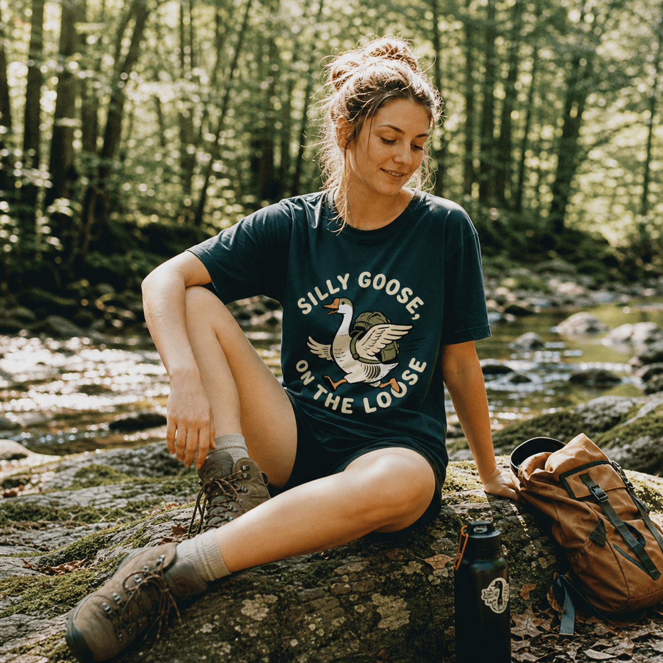 Woman relaxing by a stream in a forest, wearing a graphic t-shirt with "SILLY GOOSE ON THE LOOSE" and a goose design.