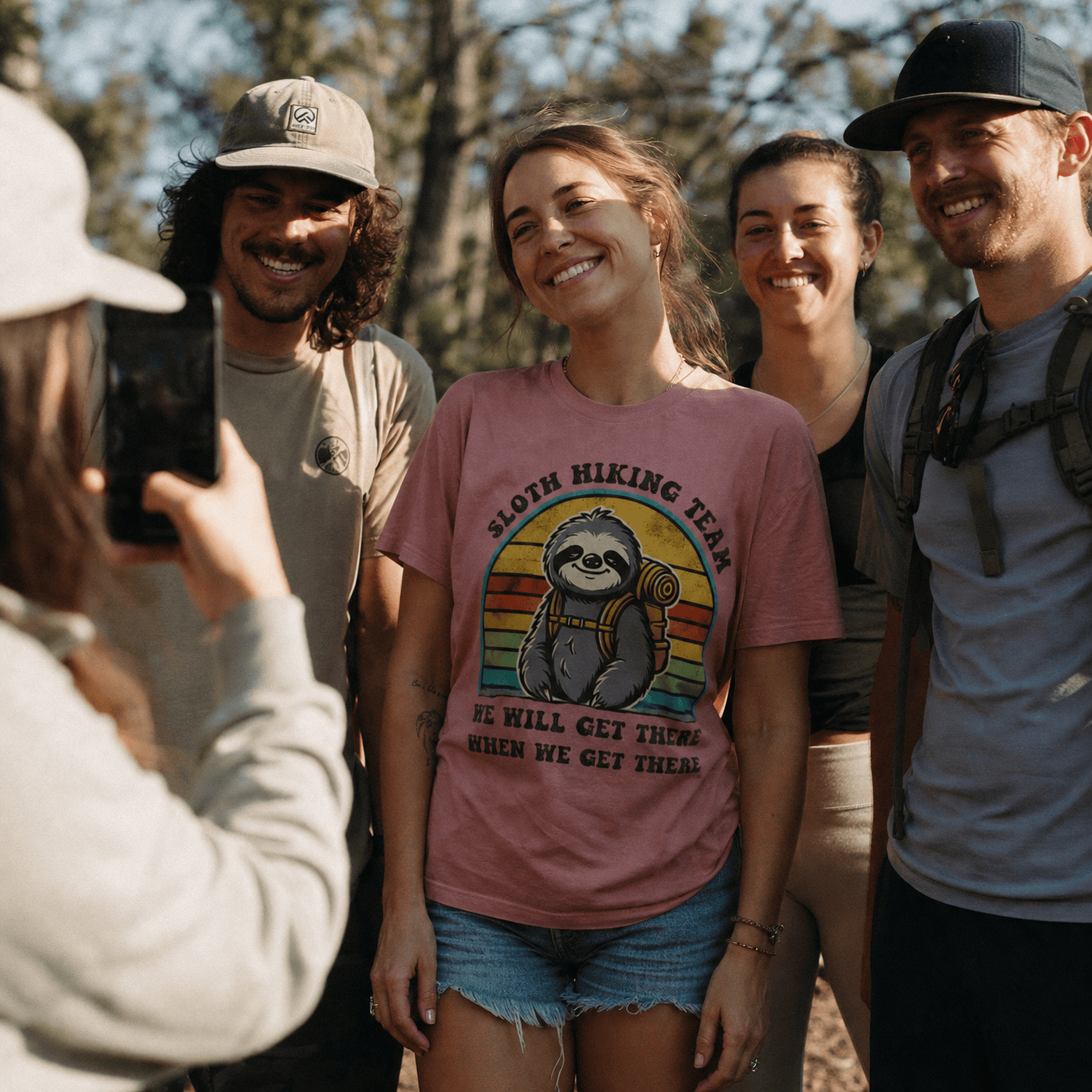 A smiling woman poses for a photo in a pink graphic t-shirt featuring the design "Sloth Hiking Team" with a sloth carrying a backpack.