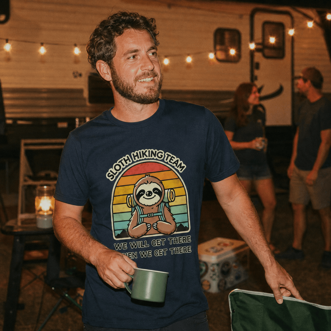 A man stands outdoors wearing a navy graphic t-shirt featuring a sloth and the words "Sloth Hiking Team" with a colorful sunset background.