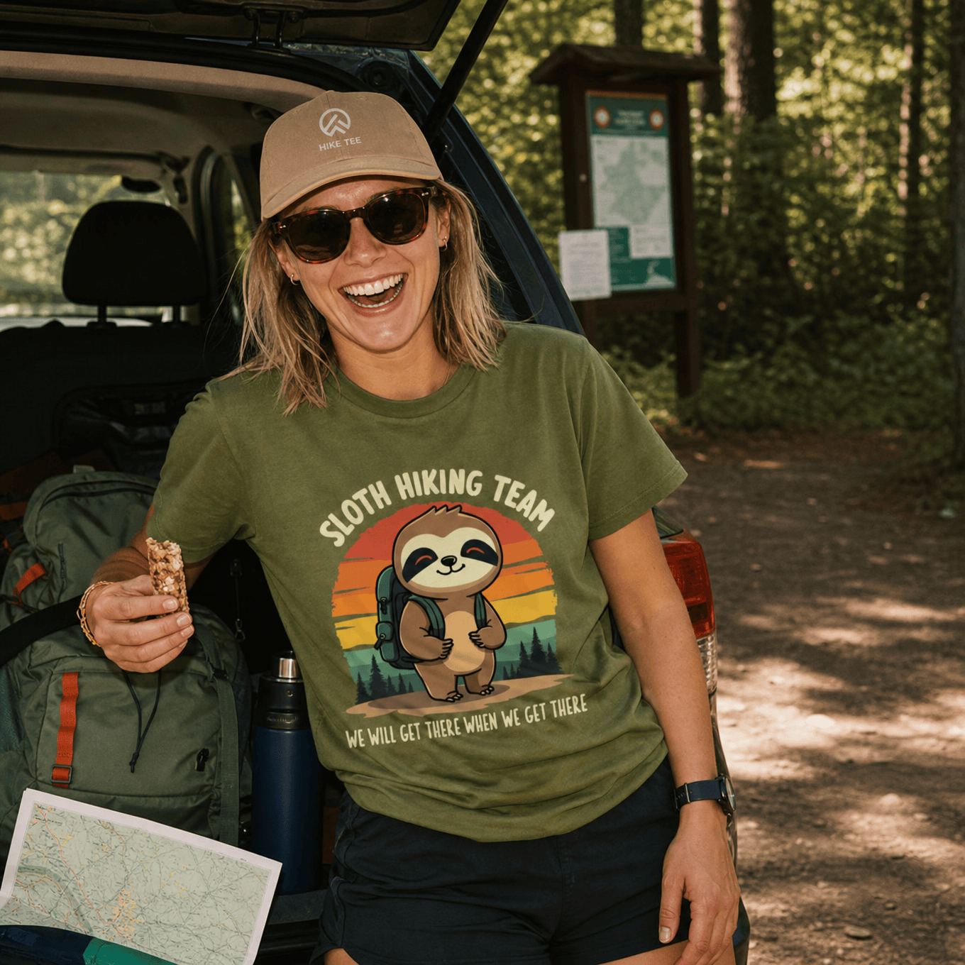 A smiling woman enjoys a snack while wearing a green graphic t-shirt featuring a sloth and the text "Sloth Hiking Team.