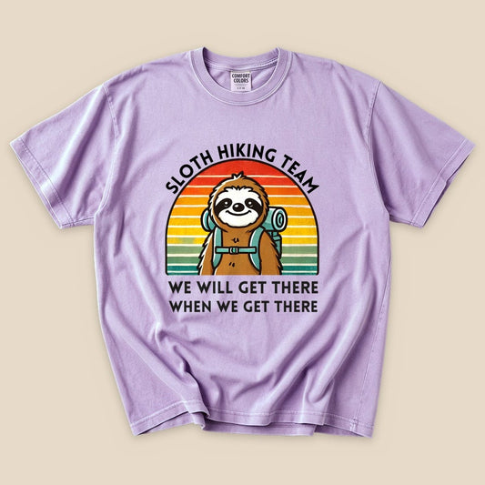 Graphic t-shirt in lavender featuring a smiling sloth with a backpack, text reads "Sloth Hiking Team" and "We Will Get There When We Get There".