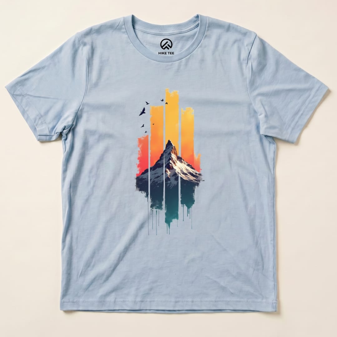 The Snow Mountain Sunset Shirt features a vibrant graphic of a mountain against a colorful sunset sky with birds, displayed on a light blue t-shirt.