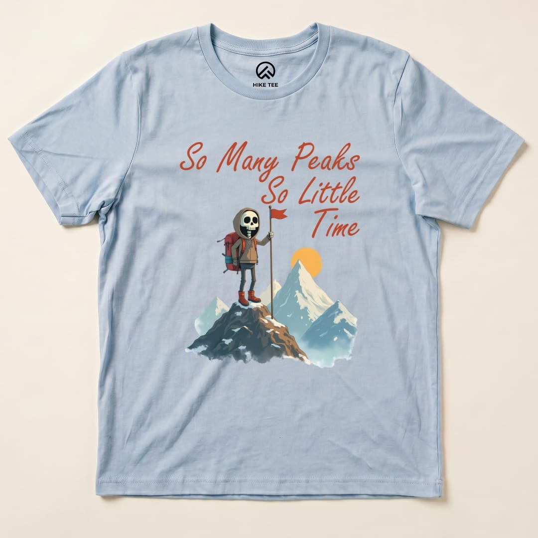Light blue graphic t-shirt featuring a cartoon skeleton hiker on a mountain with the phrase "So Many Peaks So Little Time.