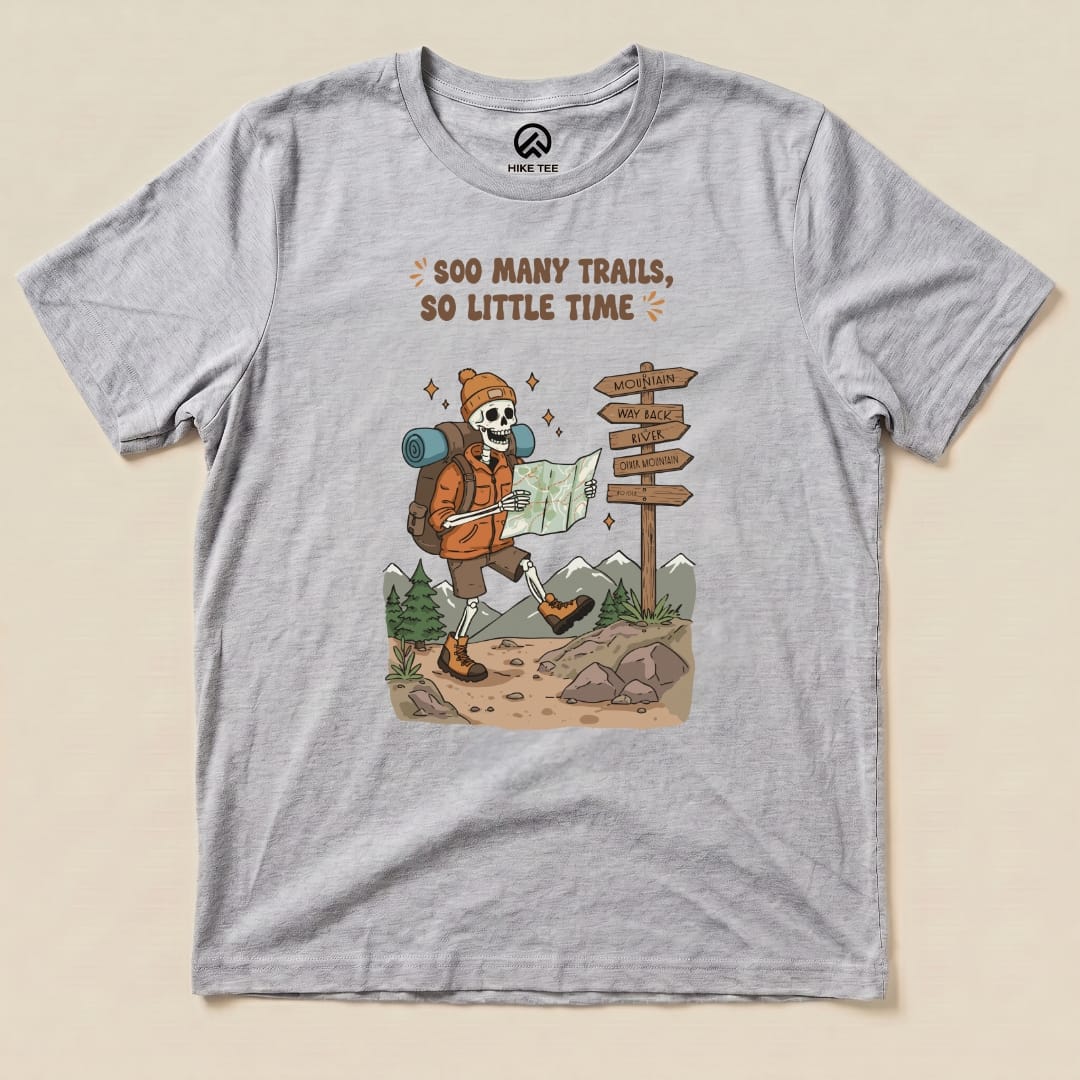 A graphic t-shirt featuring a skeleton hiker with a map, surrounded by trails and signs, captioned "So Many Trails, So Little Time.