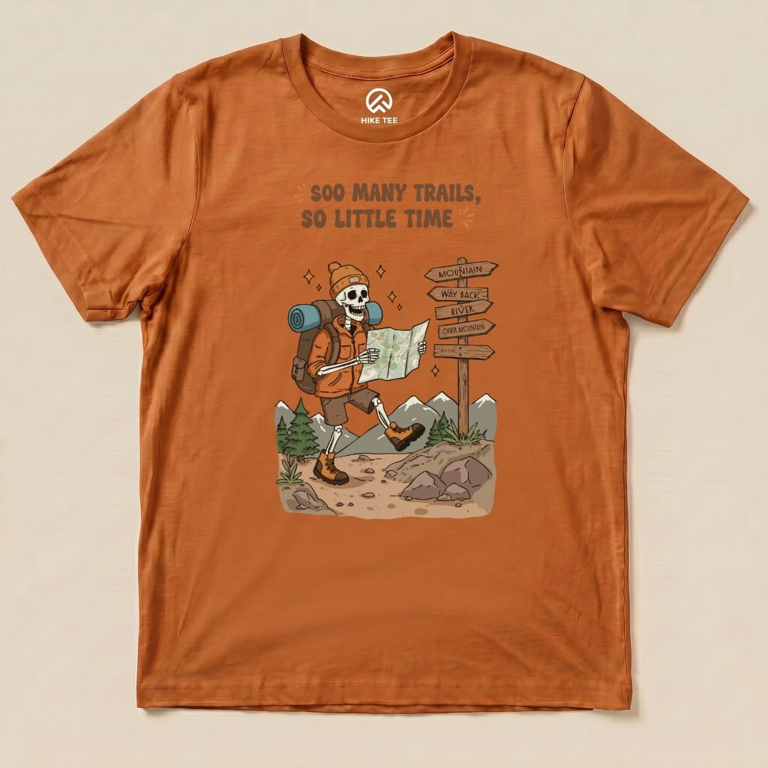 A graphic t-shirt featuring a playful skeleton hiker with the slogan "So Many Trails, So Little Time" exploring a trail map.