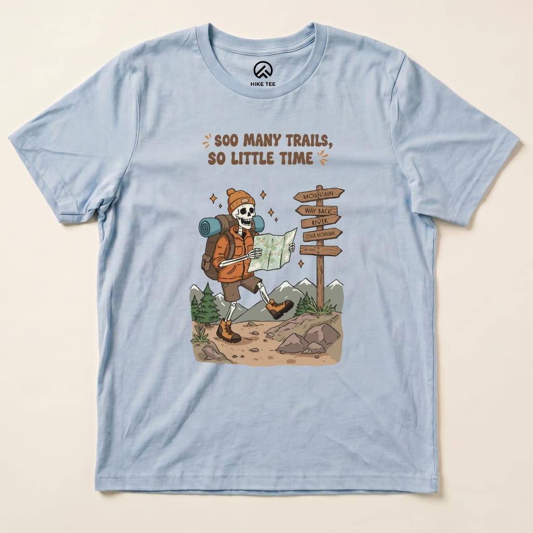 Graphic t-shirt featuring a cartoon skeleton hiking with a map and the slogan "So Many Trails, So Little Time" printed above.