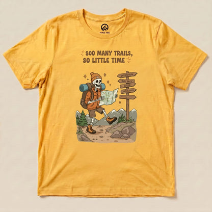 A graphic t-shirt in vibrant yellow featuring a cartoon skeleton hiker with the phrase "So Many Trails, So Little Time" and a map.