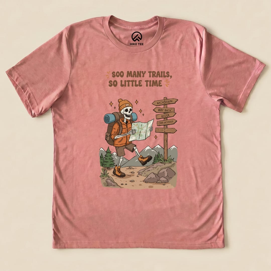 A graphic t-shirt in dusty rose featuring a playful skeleton hiker with a map and the words "So Many Trails, So Little Time.