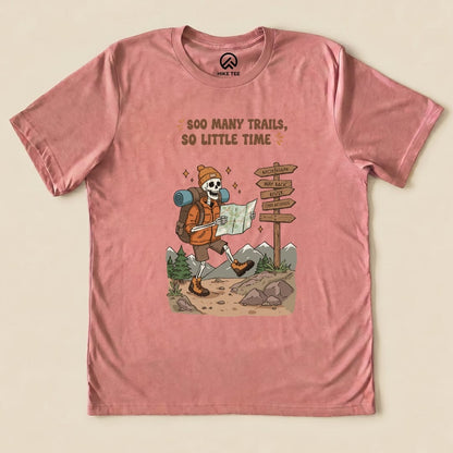 A graphic t-shirt in dusty rose featuring a playful skeleton hiker with a map and the words "So Many Trails, So Little Time.