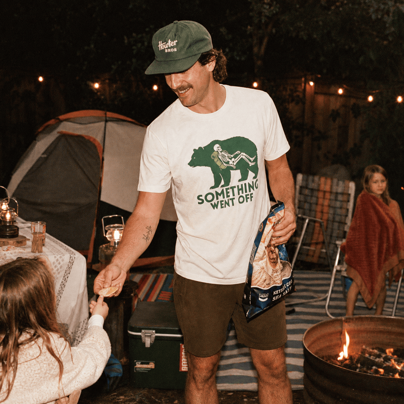 Man in a camp setting wearing a graphic t-shirt featuring a skeleton and a bear with "Something Went Off" slogan.