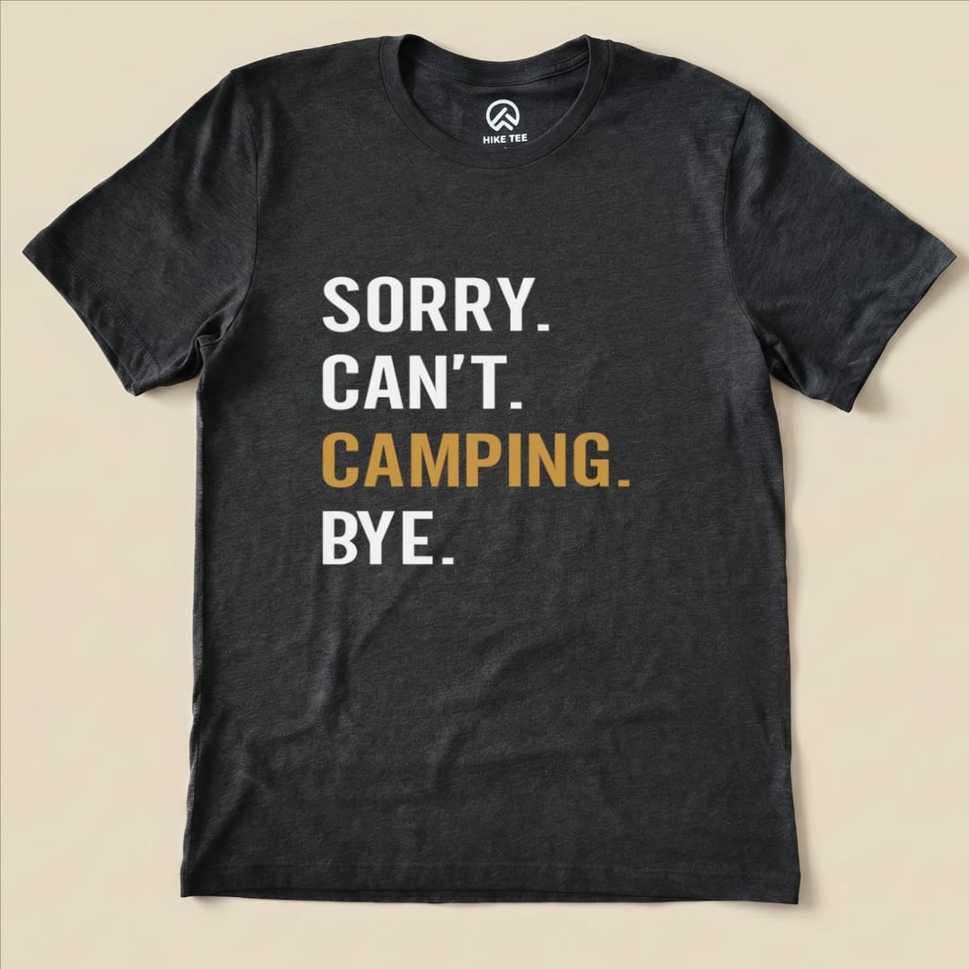 A flat lay of a charcoal graphic t-shirt featuring the text "Sorry. Can't. Camping. Bye." in bold white and gold lettering.