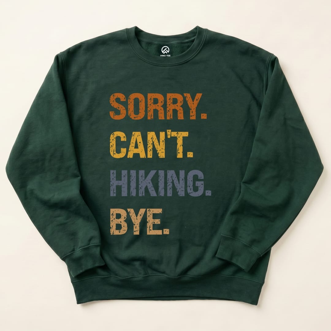 A flat lay of the "Sorry Can't Hiking Bye Sweatshirt," featuring bold text in orange, yellow, gray, and dark blue on a dark green background.