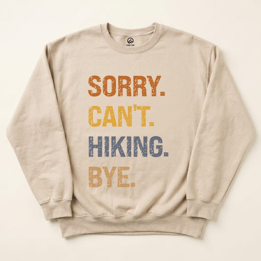 A beige sweatshirt featuring the playful text "Sorry. Can't. Hiking. Bye." in bold, colorful letters, showcasing a graphic t-shirt design.