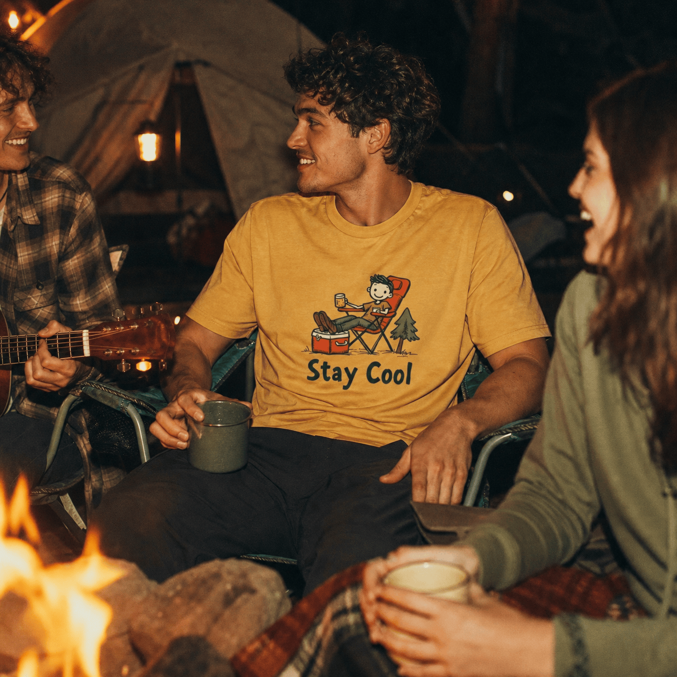 A man sits by a campfire wearing a yellow graphic t-shirt with a relaxed character design and the words "Stay Cool.