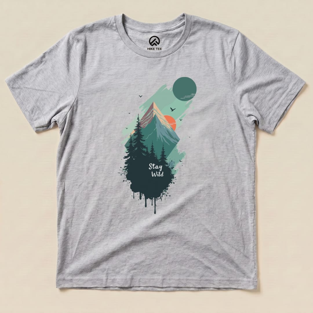 A graphic t-shirt featuring a mountainous landscape, with the words "Stay Wild" prominently displayed beneath trees and a rising sun.