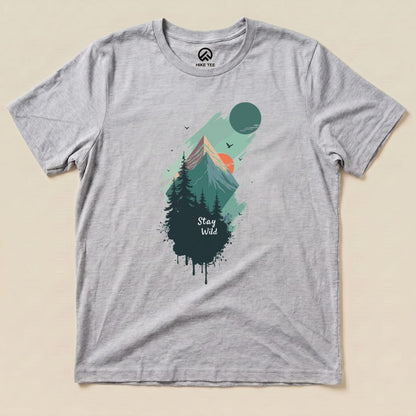 A graphic t-shirt featuring a mountainous landscape, with the words "Stay Wild" prominently displayed beneath trees and a rising sun.