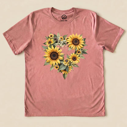 A graphic t-shirt in pink featuring a heart shape made of vibrant sunflowers and green leaves, titled "Sunflowers Heart Shirt.