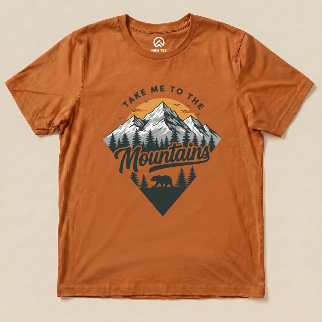 The "Take Me To The Mountains Shirt" features a stylized graphic of mountains, trees, and a bear against a warm orange background.