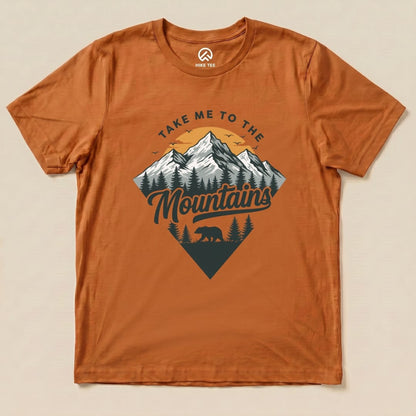 The "Take Me To The Mountains Shirt" features a stylized graphic of mountains, trees, and a bear against a warm orange background.