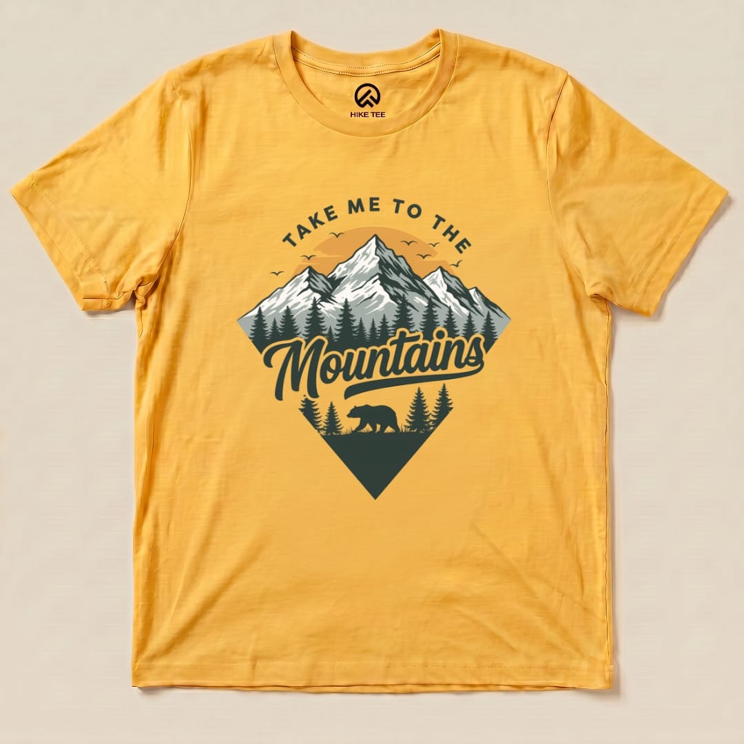 A graphic t-shirt in vibrant yellow featuring the slogan "Take Me To The Mountains" and a design of mountains, trees, and a bear.