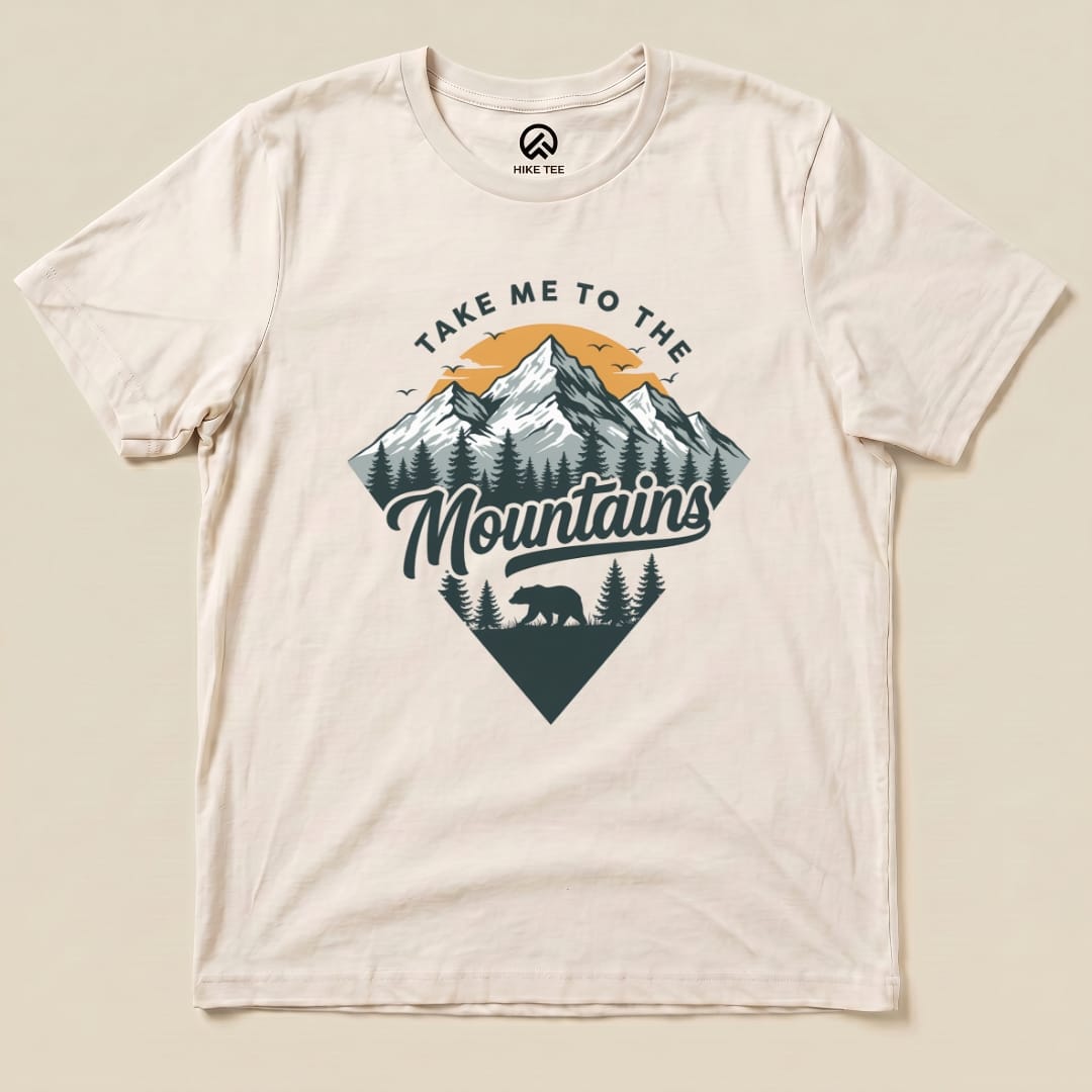 A graphic t-shirt featuring the slogan "Take Me To The Mountains" along with a mountain scene and a silhouette of a bear.