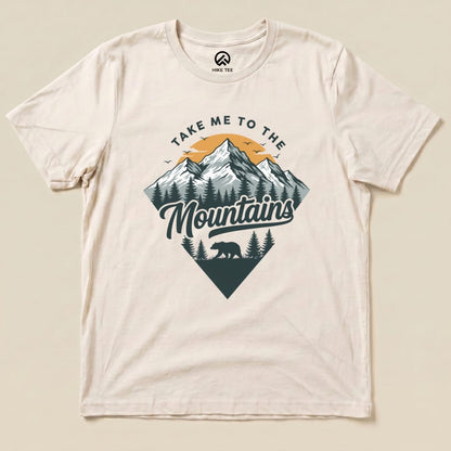 A graphic t-shirt featuring the slogan "Take Me To The Mountains" along with a mountain scene and a silhouette of a bear.