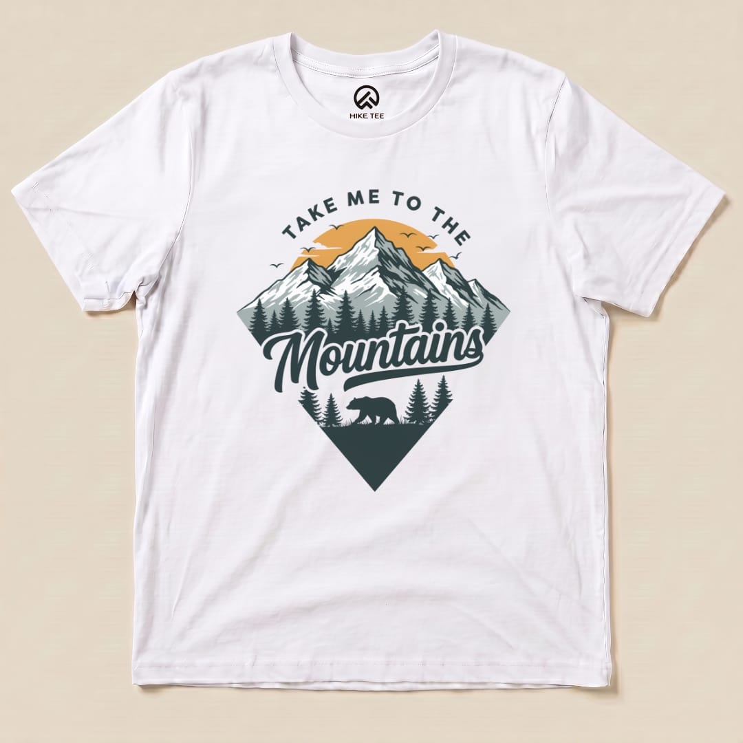 A flat white graphic t-shirt featuring the slogan "Take Me To The Mountains" alongside a mountain and bear design.