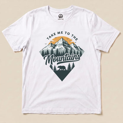 A flat white graphic t-shirt featuring the slogan "Take Me To The Mountains" alongside a mountain and bear design.