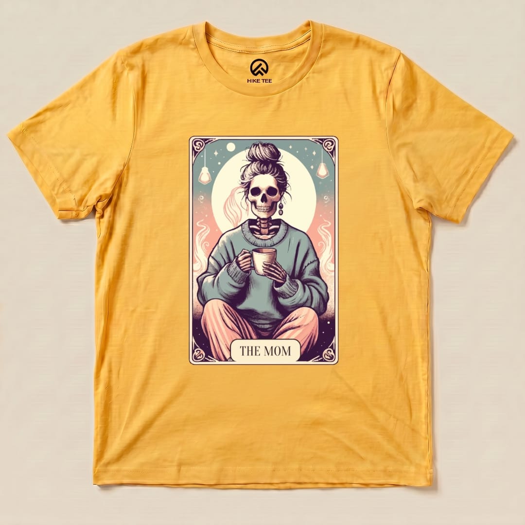A yellow graphic t-shirt features a design of a skeleton in a sweater holding a cup, labeled "THE MOM," identified as a Tarrot Mom Shirt.