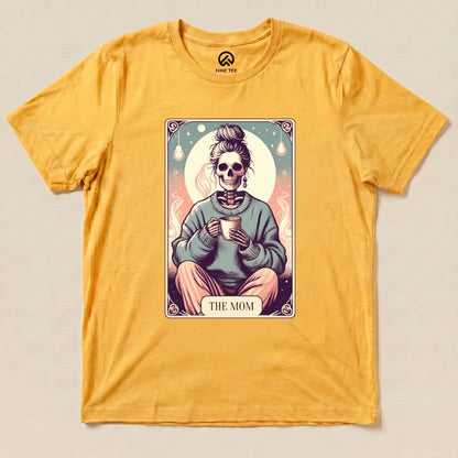 A yellow graphic t-shirt features a design of a skeleton in a sweater holding a cup, labeled "THE MOM," identified as a Tarrot Mom Shirt.