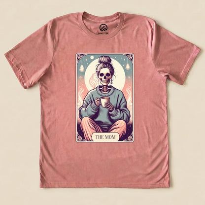 The Tarrot Mom Shirt features a graphic of a skeleton in a cozy sweater holding a cup, framed by whimsical elements.
