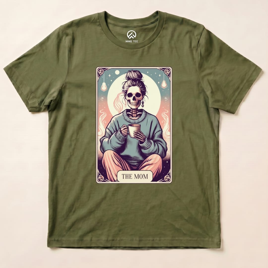 A graphic t-shirt featuring a skeleton wearing a cozy sweater, holding a mug, with the words "THE MOM" below the design, called the Tarrot Mom Shirt.