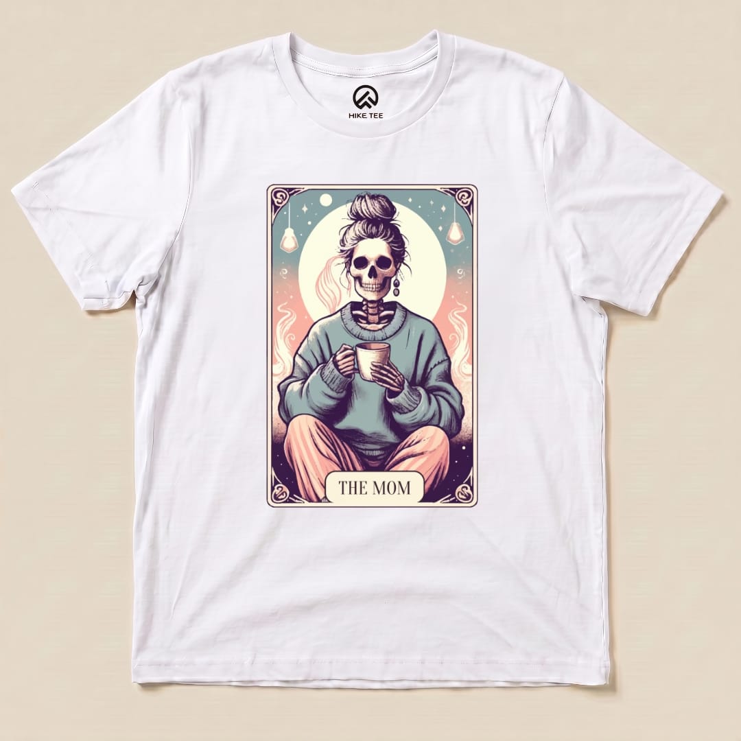 A white graphic t-shirt featuring a whimsical illustration of a skeleton with a mug, labeled "THE MOM," labeled as the Tarrot Mom Shirt.
