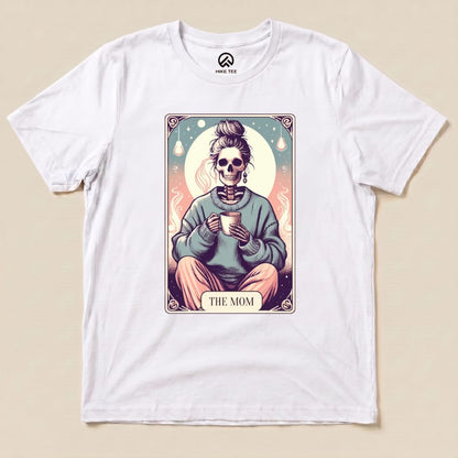 A white graphic t-shirt featuring a whimsical illustration of a skeleton with a mug, labeled "THE MOM," labeled as the Tarrot Mom Shirt.