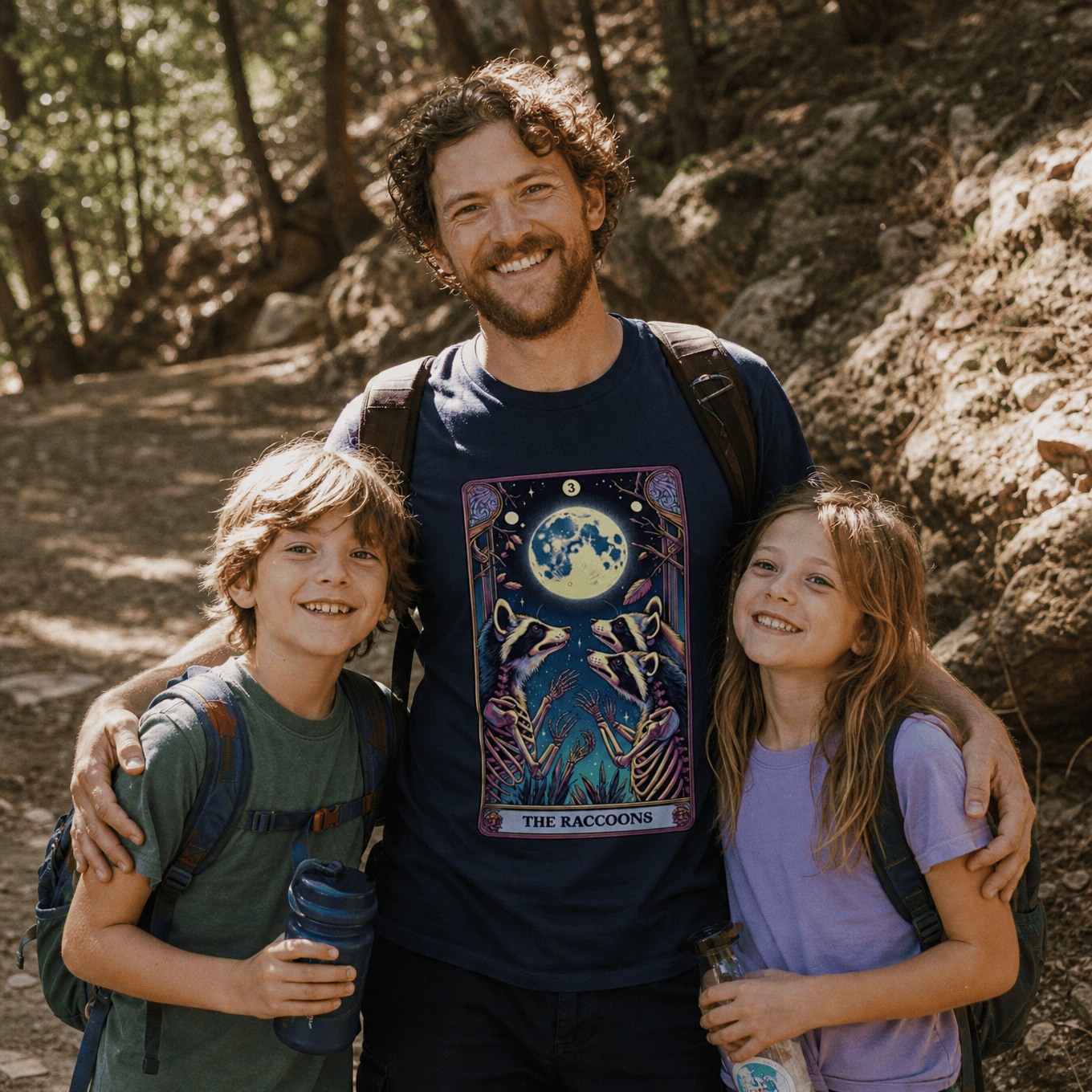 A man smiles with two children on a hiking trail, wearing a graphic t-shirt featuring the "Tarrot Raccoon Shirt" design.