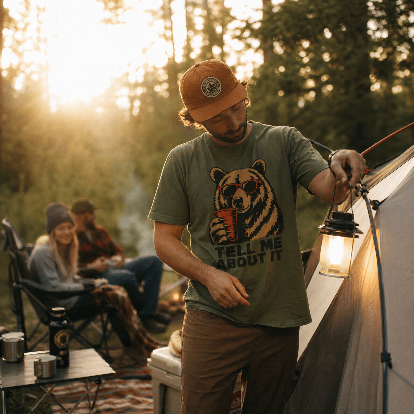 A man outdoors at a campsite wears a graphic t-shirt featuring a bear in sunglasses and the phrase "TELL ME ABOUT IT.