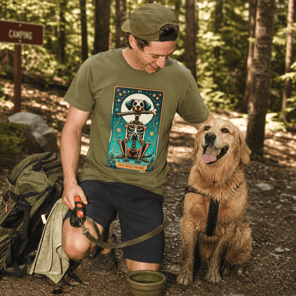 A man kneels in the woods wearing a graphic t-shirt featuring the "Th Best Friend Tarot Card Dog Shirt" design with a playful skeleton.