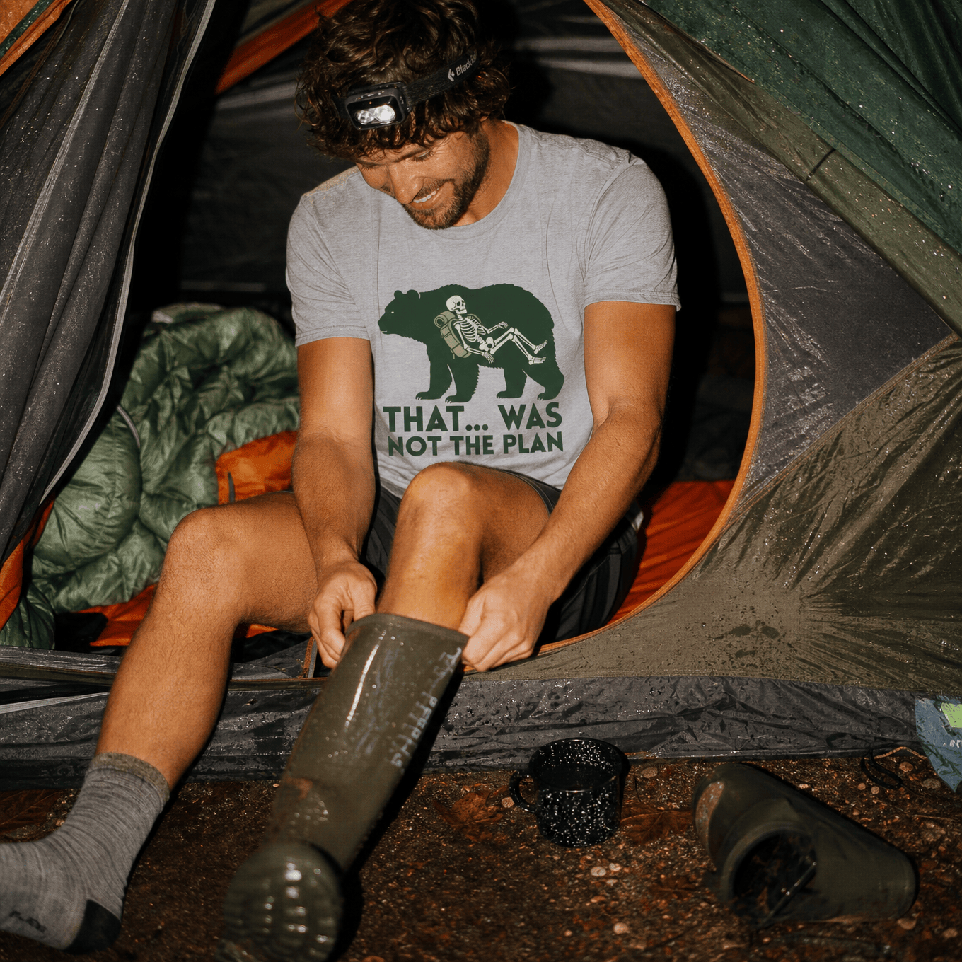 A smiling man in a tent puts on boots, wearing a gray graphic t-shirt featuring a bear and the slogan "That... Was Not The Pl