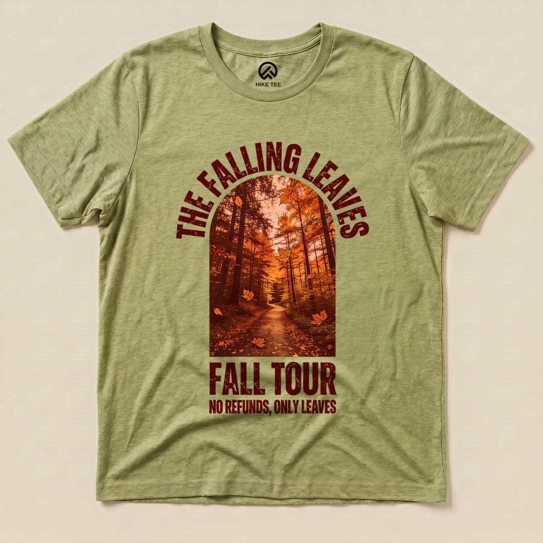 The Falling Leaves Fall Tour Shirt features a vibrant autumn scene with a forest pathway and falling leaves, printed on a green graphic t-shirt.