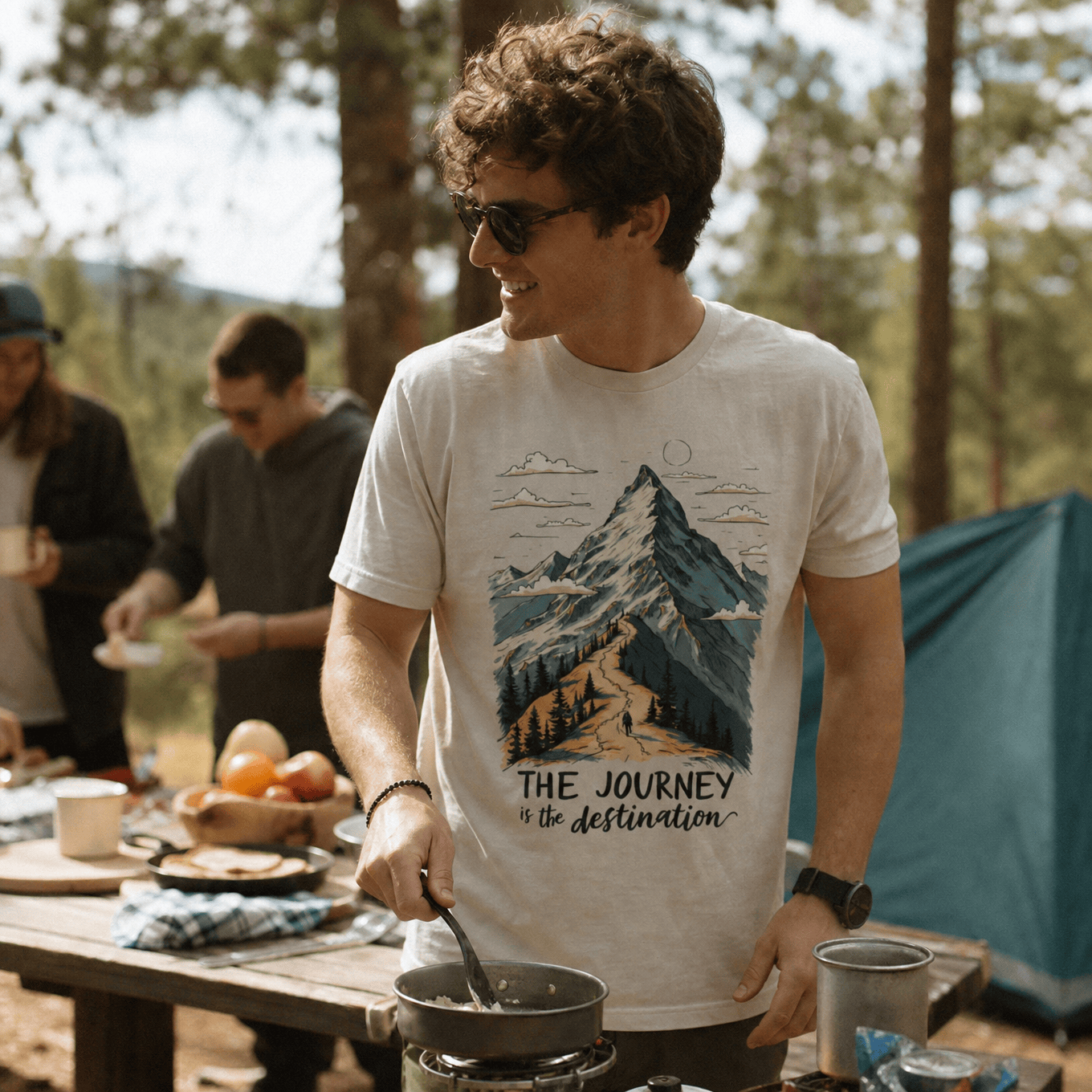 A man is cooking outdoors while wearing "The Journey Shirt," featuring a graphic of a mountain with the slogan "THE JOURNEY is the destination.