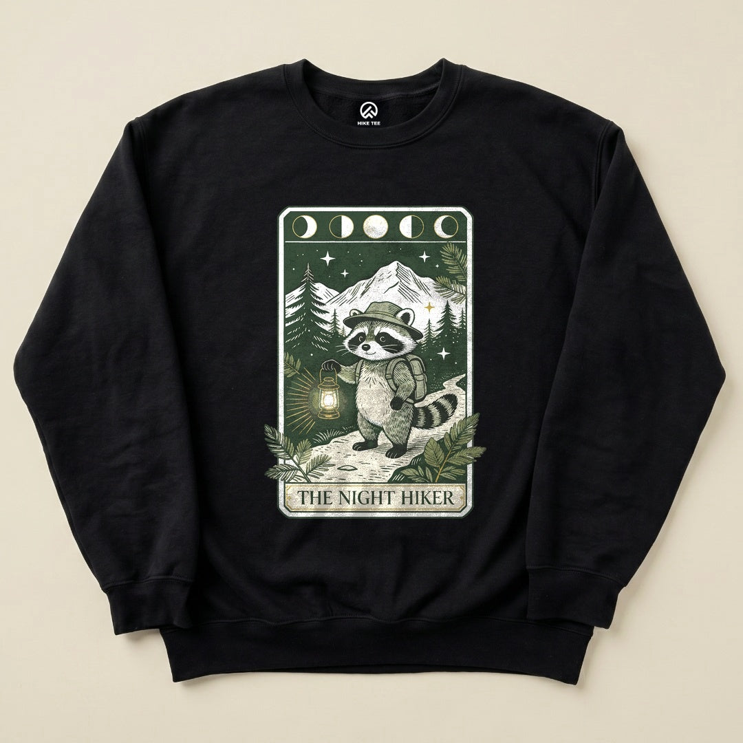 A black sweatshirt featuring a raccoon character with a lantern, surrounded by mountains and trees, labeled "THE NIGHT HIKER.
