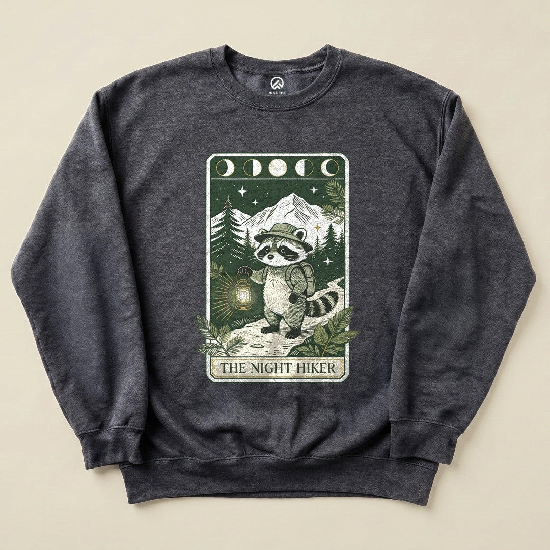 A graphic sweatshirt featuring a raccoon carrying a lantern on a trail, surrounded by trees and mountains, labeled "THE NIGHT HIKER.