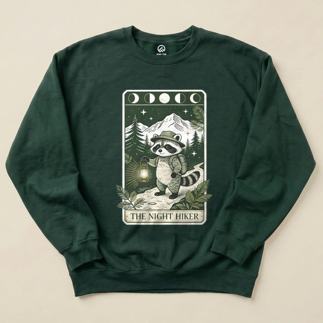 The Night Hiker Sweatshirt features a raccoon character holding a lantern, surrounded by mountains and trees in a vintage graphic design.