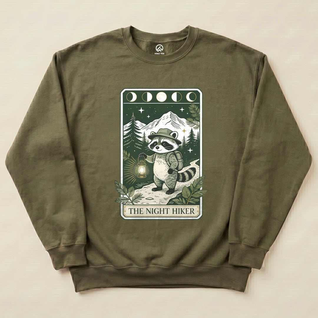 The Night Hiker Sweatshirt features a playful raccoon holding a lantern, set against a backdrop of mountains and trees.