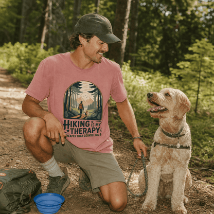 A man kneels on a trail with his dog, wearing a pink graphic t-shirt that reads "Hiking is my therapy, cheaper than counseling.