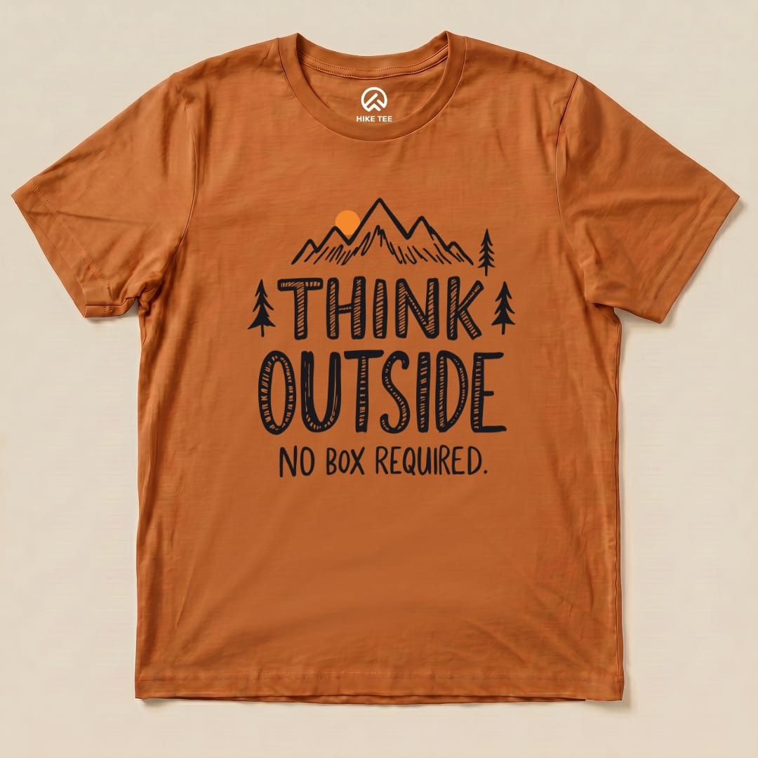 A flat graphic t-shirt in a rust color featuring the slogan "THINK OUTSIDE NO BOX REQUIRED" with mountain and tree illustrations.