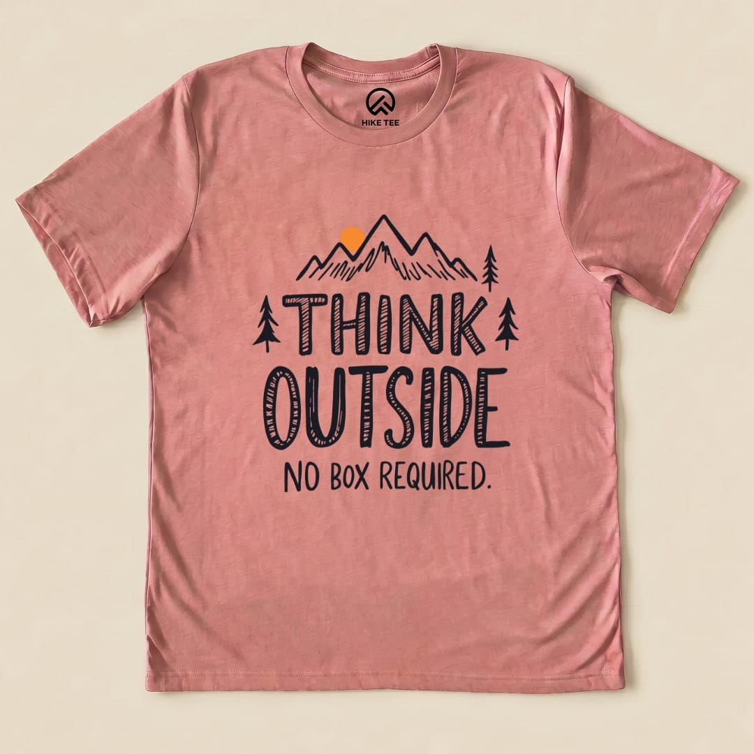 A flat lay of a pink graphic t-shirt featuring the phrase "THINK OUTSIDE" above an illustration of mountains and trees, with "NO BOX REQUIRED.