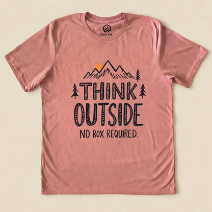 A flat lay of a pink graphic t-shirt featuring the phrase "THINK OUTSIDE" above an illustration of mountains and trees, with "NO BOX REQUIRED.